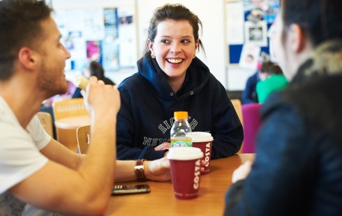 Female student laughing and sat drinking coffee with two male students.