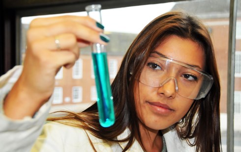 Close up image of female wearing plastic protective goggles, white lab coat and holding a test tube with light blue liquid in.