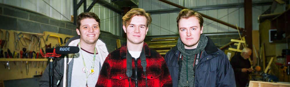 Three male students smiling with camera equipment in a construction warehouse
