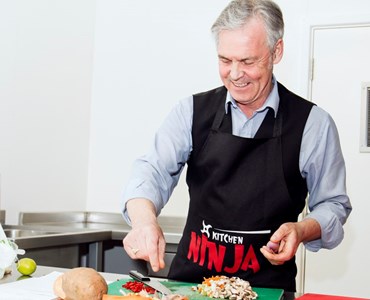 Grey haired man wearing black apron and smiling while preparing food on a green chopping board.