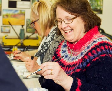 Mature female student smiling and holding a paintbrush while sat in a classroom.