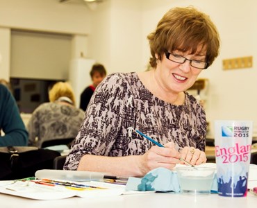Mature female student smiling while sat at a desk painting with water colours.