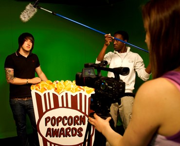 3 students in a green screen room, one student is behind the camera, one holding a microphone and the other student is stood behind a large popcorn cardboard cut
