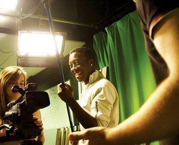 3 students in a green screen studio, one student is stood behind a large recording camera and one is holding a recording microphone