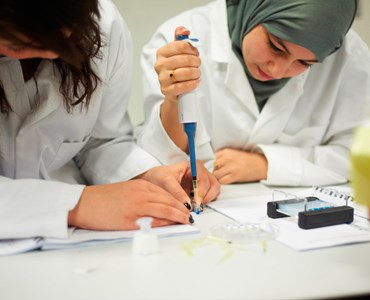 2 female students in science laboratory coats sat at a desk together concentrating on a science experiment