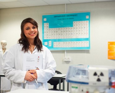A female student in a laboratory coat holding a workbook stood in a science laboratory smiling at the camera