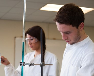 A female and male student in a science laboratory in protective lab coats,  the female student is wearing protective goggles and is holding a test tube with blue liquid