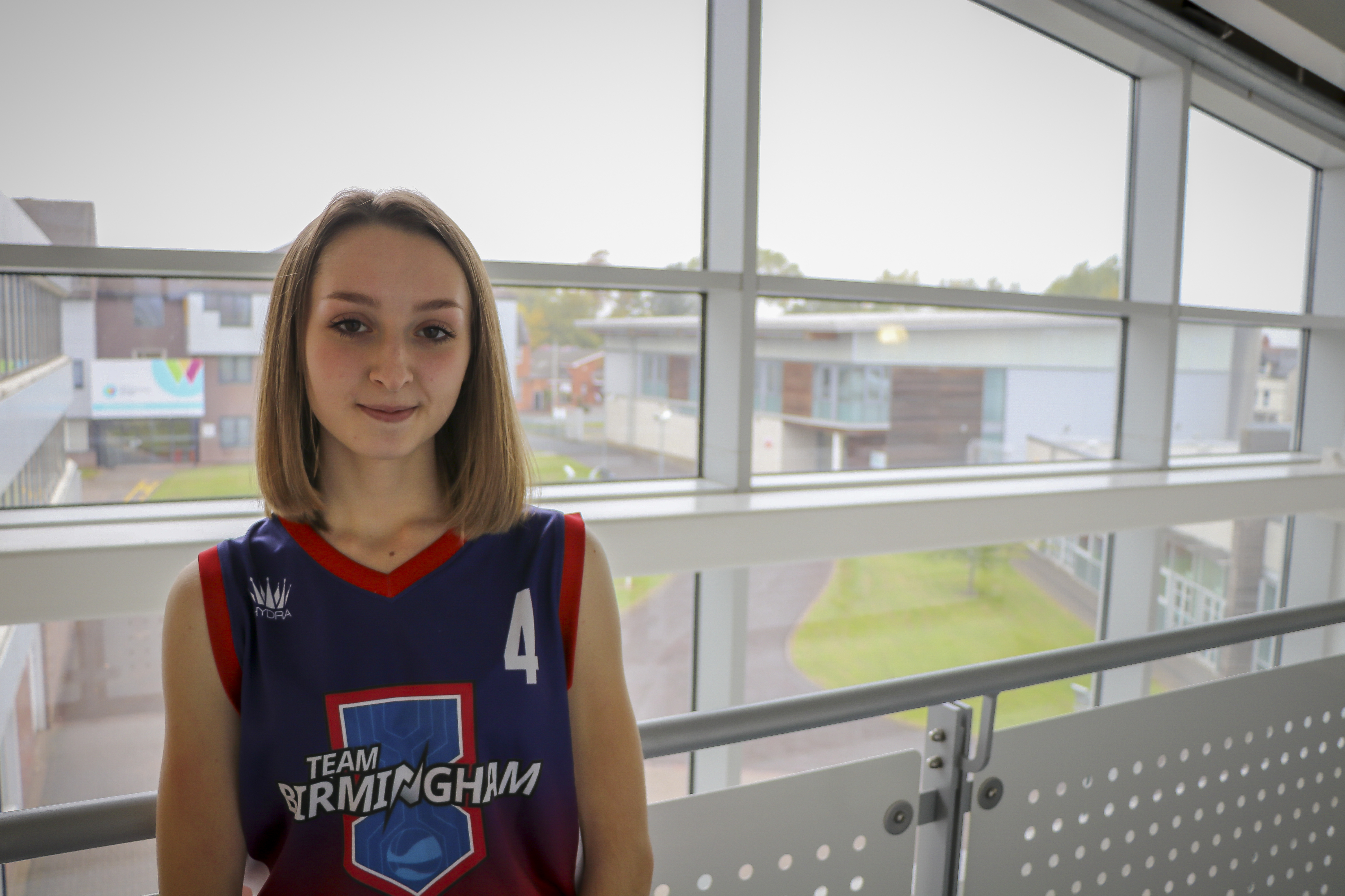 Female student wearing a Team Birmingham navy and red sleeveless sports jersey in front of a window