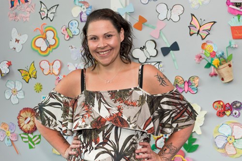 Female stood smiling with her hands on her hips in front of a grey wall with colourful paper butterflies and flowers on