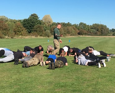 Group of male and female students holding the plank position in a large circle in the middle of a field, with a marine holding a stop watch in the middle.