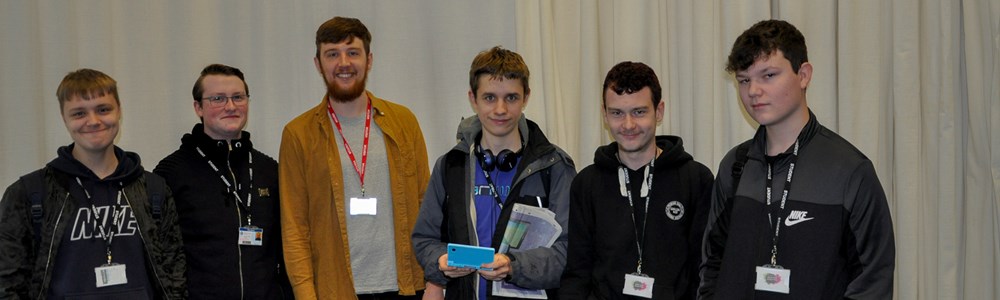 Group of male students stood smiling in front of large white drapes. Student in the middle holding a blue, handheld games device. 