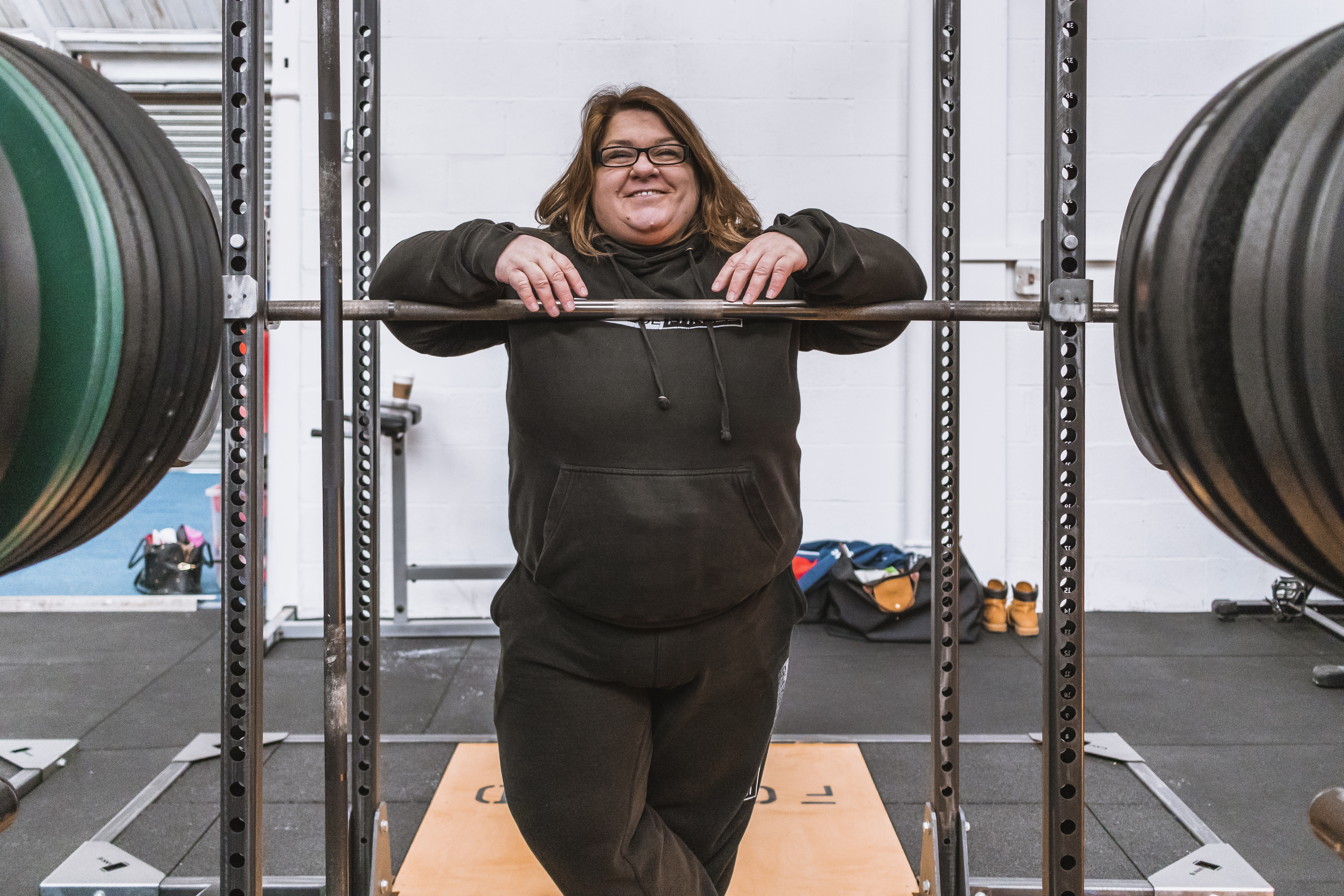 Image of female in black hoodie smiling and resting her arms on a squat bar in a gym