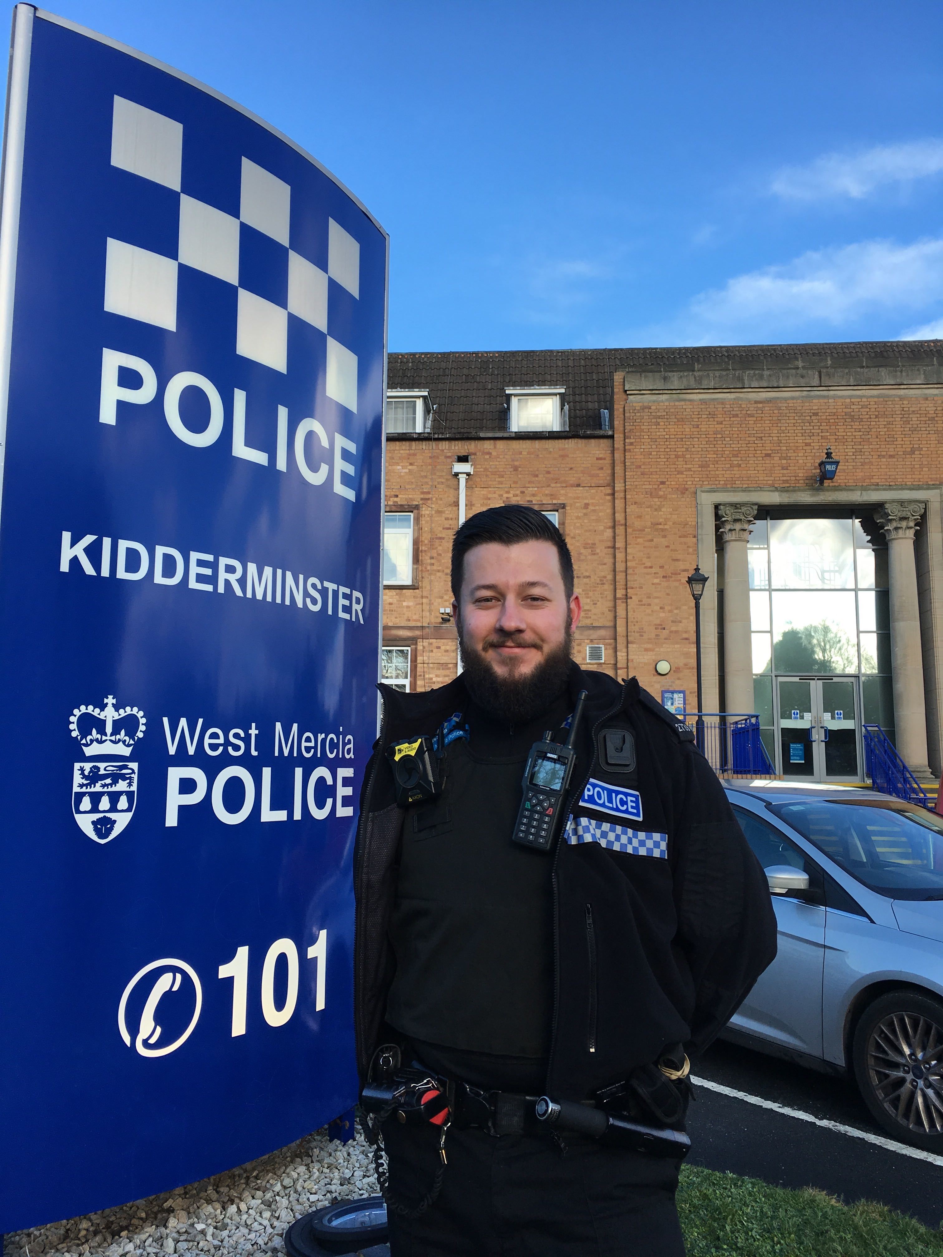 Male police officer with beard stood in uniform and smiling in front of the West Mercia Police Kidderminster sign