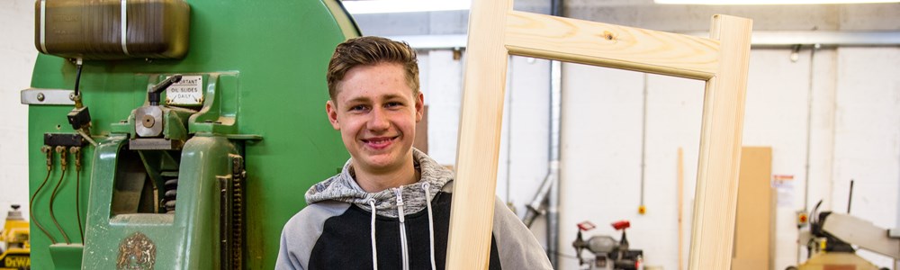 Smiling male student in front of green machine while holding a large, rectangular wooden item