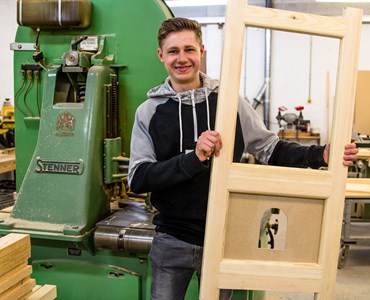 Smiling male student in front of green machine while holding a large, rectangular wooden item