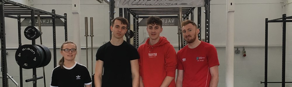 4 male and female students stood smiling on a black and wooden weightlifting platform in a gym.