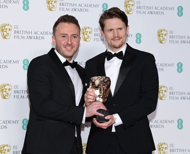 2 male adults in black tuxedos holding a Bafta award together while stood in front of a backdrop with Bafta logos on.