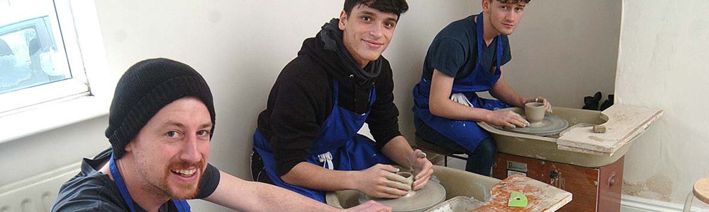 3 smiling male students in blue aprons molding clay pottery.