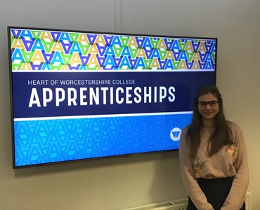 Female student stood in front of large TV with the Heart of Worcestershire College Apprenticeships logo on