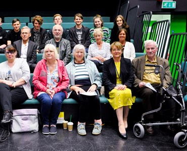 Group of senior adults and college students sat on green theatre benches in a black TV studio