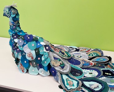 Large blue peacock made of lots of pieces of fabric and placed on a white table in front of a green wall.