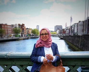 Female wearing a pink hijab and navy blazer stood holding a large brown leather handbag in front of a green bridge. Water and city can be seen in the background.