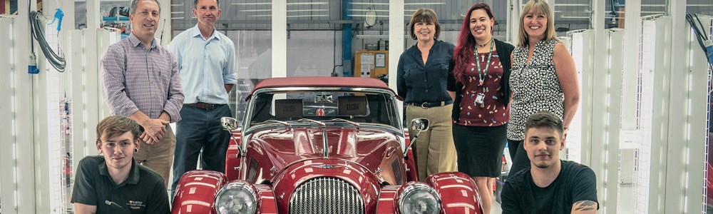 Males and females stood around a cherry red vintage car in a brightly lit white garage