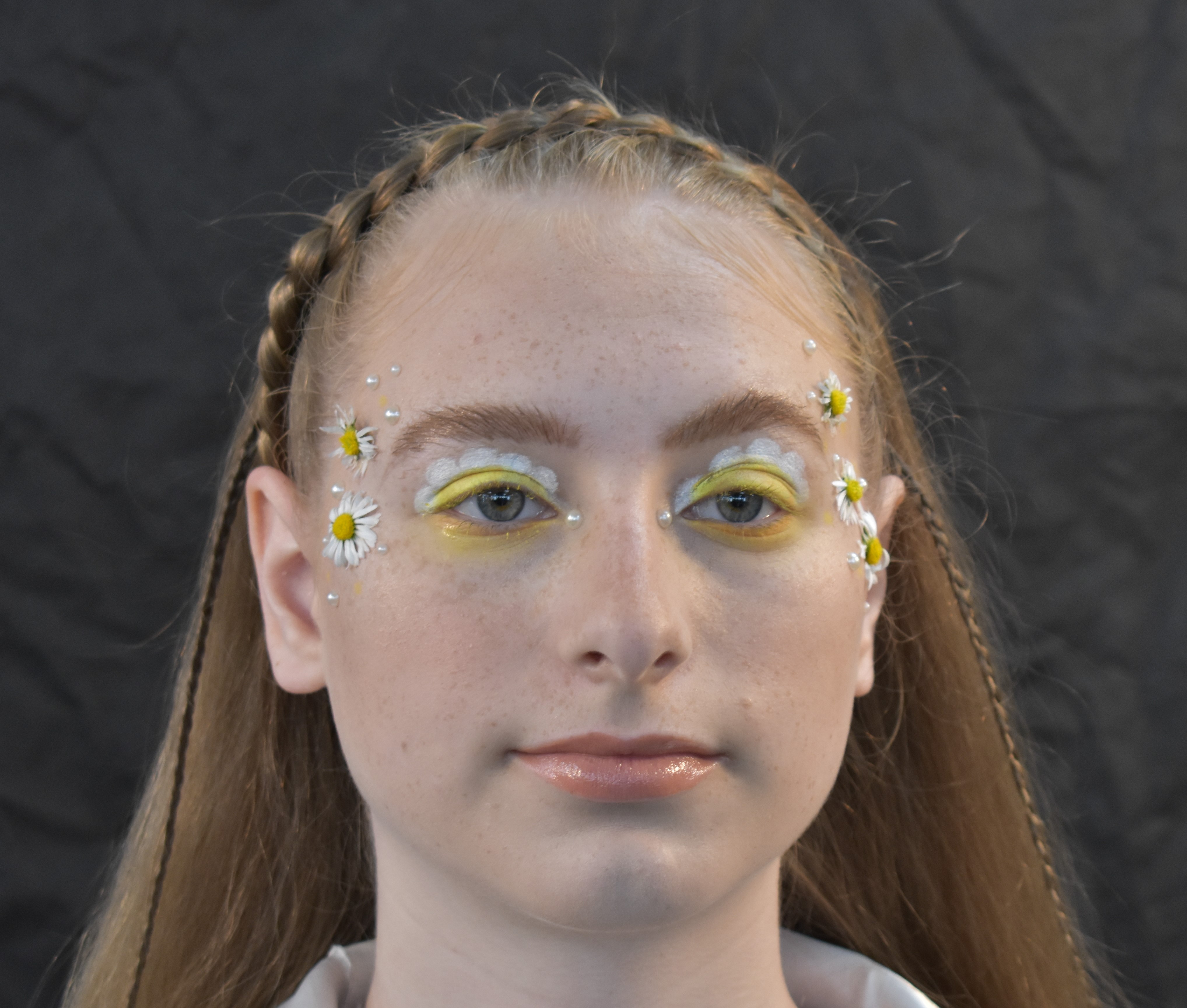 Close up facial picture of female with yellow and white flowery eye makeup, daisies attached to her temples and with small braid across the top of her head.