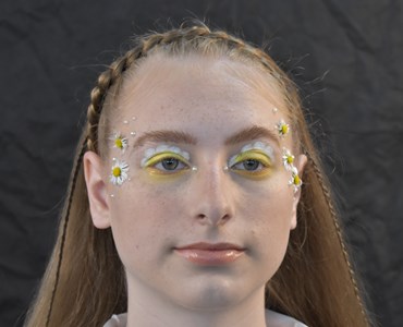 Close up facial picture of female with yellow and white flowery eye makeup, daisies attached to her temples and with small braid across the top of her head.