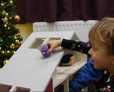 Male child in pajamas playing with a small purple toy and large wooden dolls house; Christmas tree is visible in the background.
