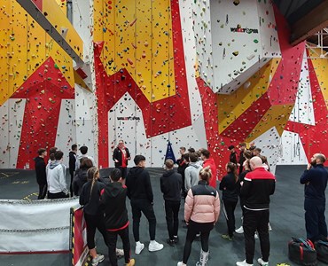 Large group of students with their back to the camera, in front of a large white, red and yellow climbing wall.