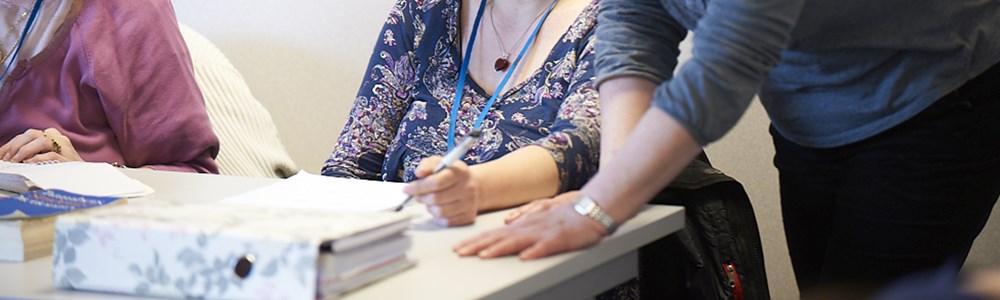 A cropped photo of two people sat at a desk writing with a person stood over one of the people