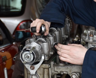 A close up of a student wearing overalls working on a car engine with a red car to the side