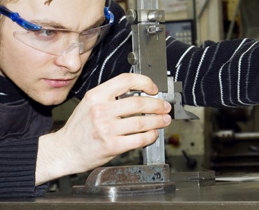 A male student wearing protective goggles in an engineering workshop looking closely at machinery