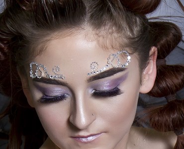 A close up of a female in artistic makeup with purple eye shadow and silver diamante swirls above the eyebrows