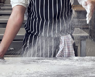 A close up a student in chef whites and striped apron applying flour to a kitchen worktop