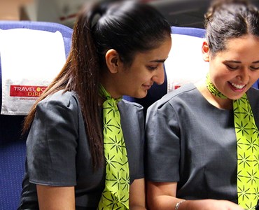 Three female travel and tourism students in air hostess uniforms and reading a magazine together are sat in the front row of mock air cabin seats