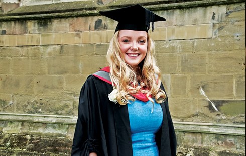 Female student smiling in graduation cap and gown in front of cathedral wall.
