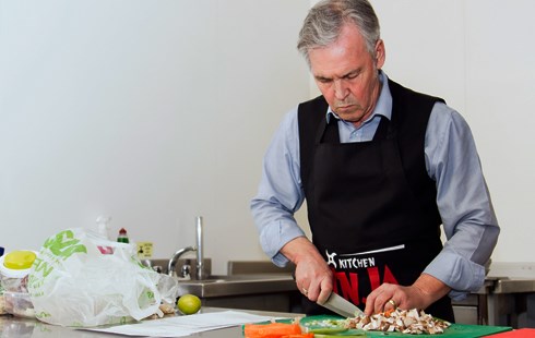 Grey haired man in black apron chopping and slicing vegetables.