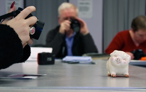Image of white piggy bank on desk with student holding a camera and taking a photo.