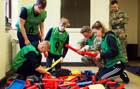 Image of 4 students in green bibs working with large plastic parts; 2 males in army uniform monitoring.