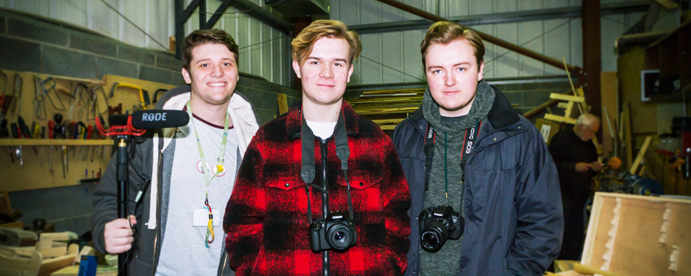 Three male students smiling with camera equipment in a construction warehouse