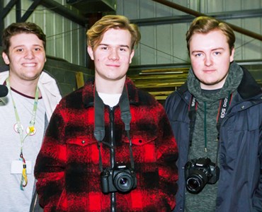 Three male students smiling with camera equipment in a construction warehouse