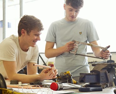Two male students smiling together at wooden desk with metal vice and hand saws.