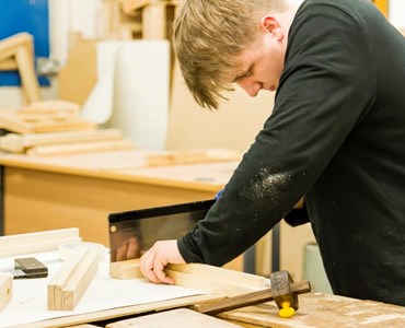 Male student using a hand saw to chop some wood at a wooden desk.