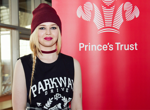 Female smiling and wearing a red beanie hat whilst stood next to a red vertical banner that reads: Prince's Trust