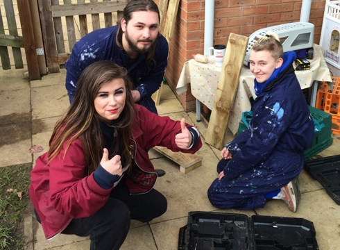 3 construction students crouching outside in a patio area. 2 males wearing blue overalls and 1 female wearing a burgundy hoodie and giving two thumbs up.