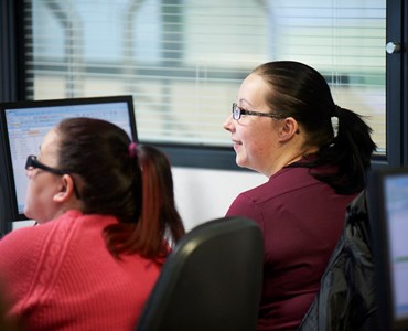 Two females smiling and working behind computers.