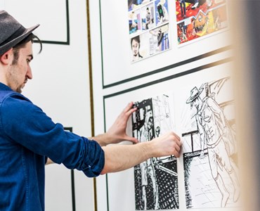 Male student in trilby hat placing comic inspired art work on a white wall.