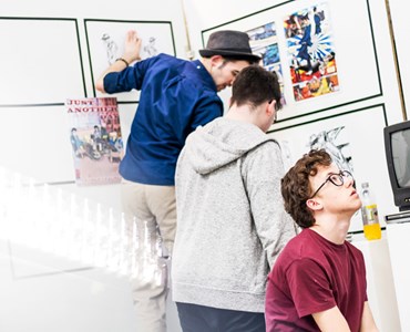 2 Male students hanging artwork on white wall; one male student looking up at another wall of art.